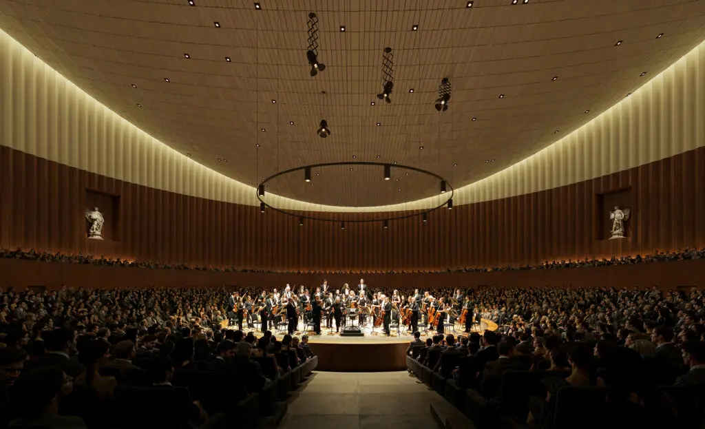 A large orchestra on a luminous stage, encircled by audience; refined wooden walls and an arching ceiling define the acoustic space.